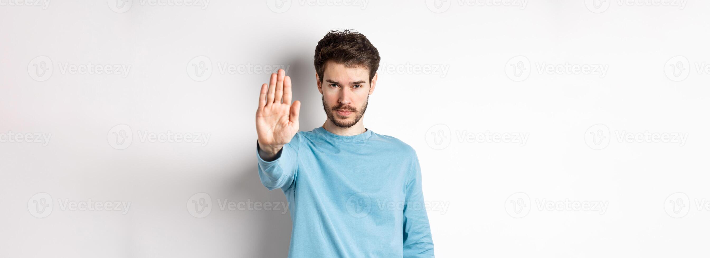 Serious young man looking confident with hand stretched out, saying stop, prohibit something bad or give warning, standing over white background photo