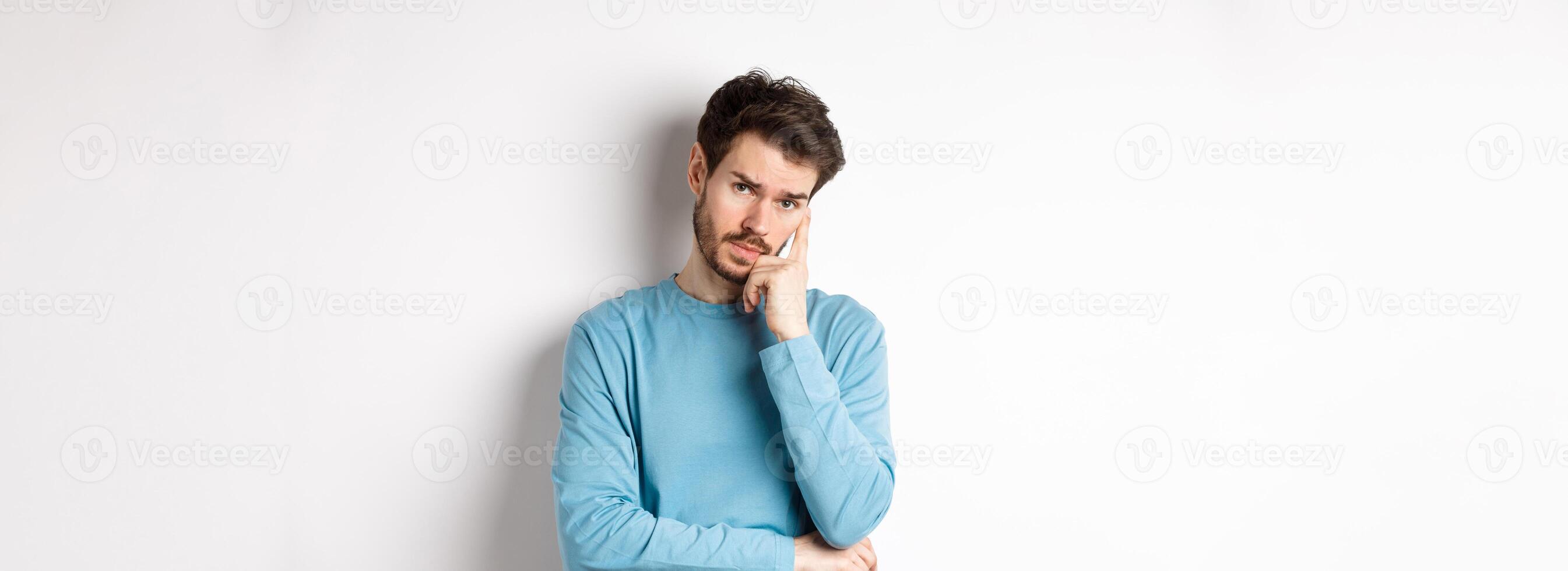 Skeptical young man listening to you with bored face, looking at camera reluctant, standing in sweatshirt over white background photo