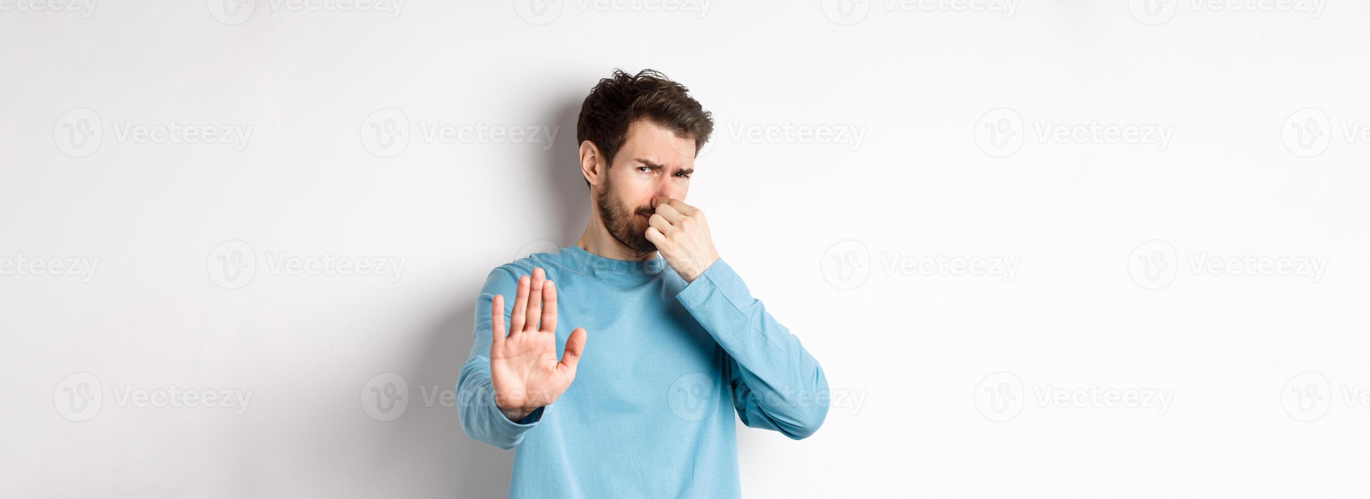 Image of caucasian man stretch out hands to stop or reject something, shut nose disgust, refusing product with bad smell, standing over white background photo