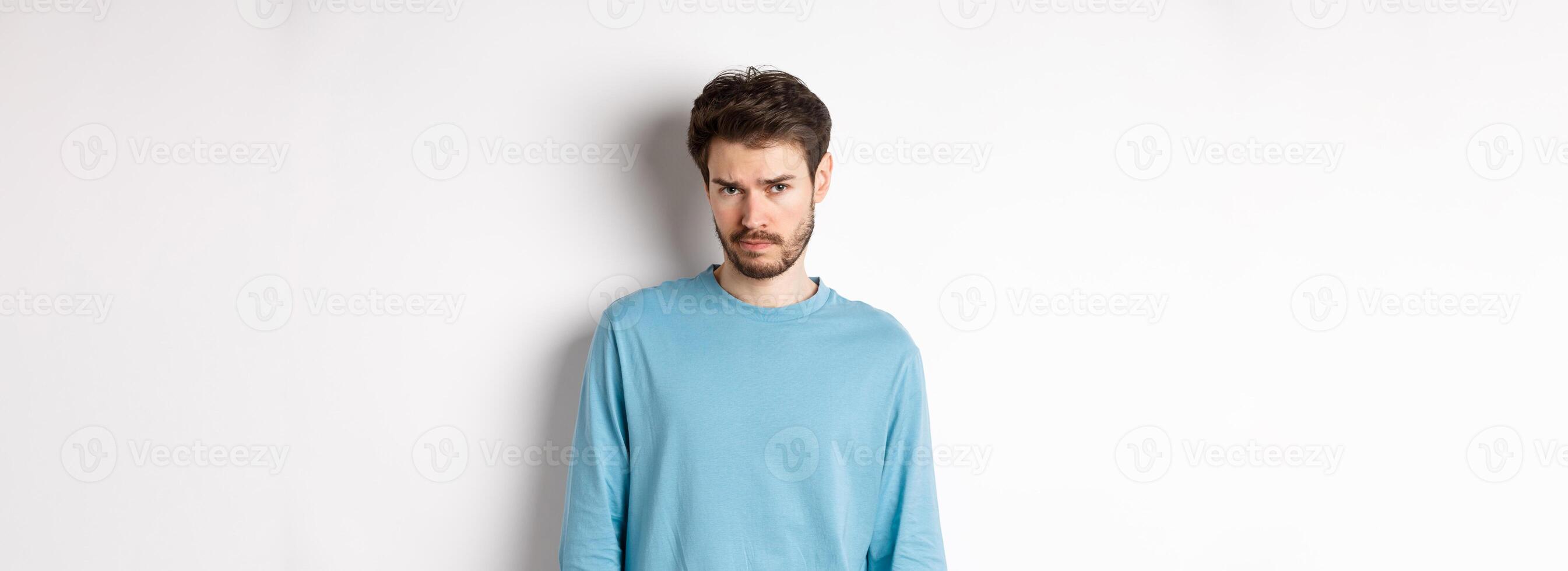 Sad young bearded man sulking, looking upset at camera, offended by someone, standing against white background photo