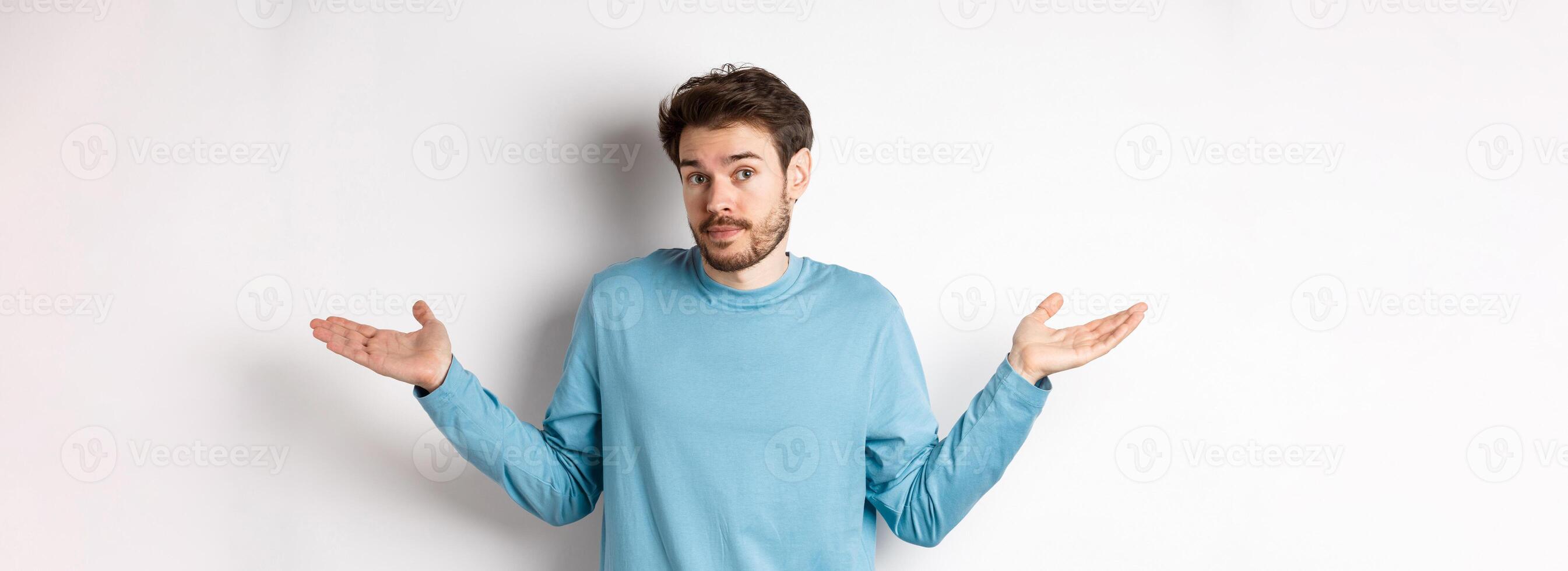 Clueless bearded guy in blue shirt, shrugging shoulders and looking unaware at camera, know nothing, standing on white background photo