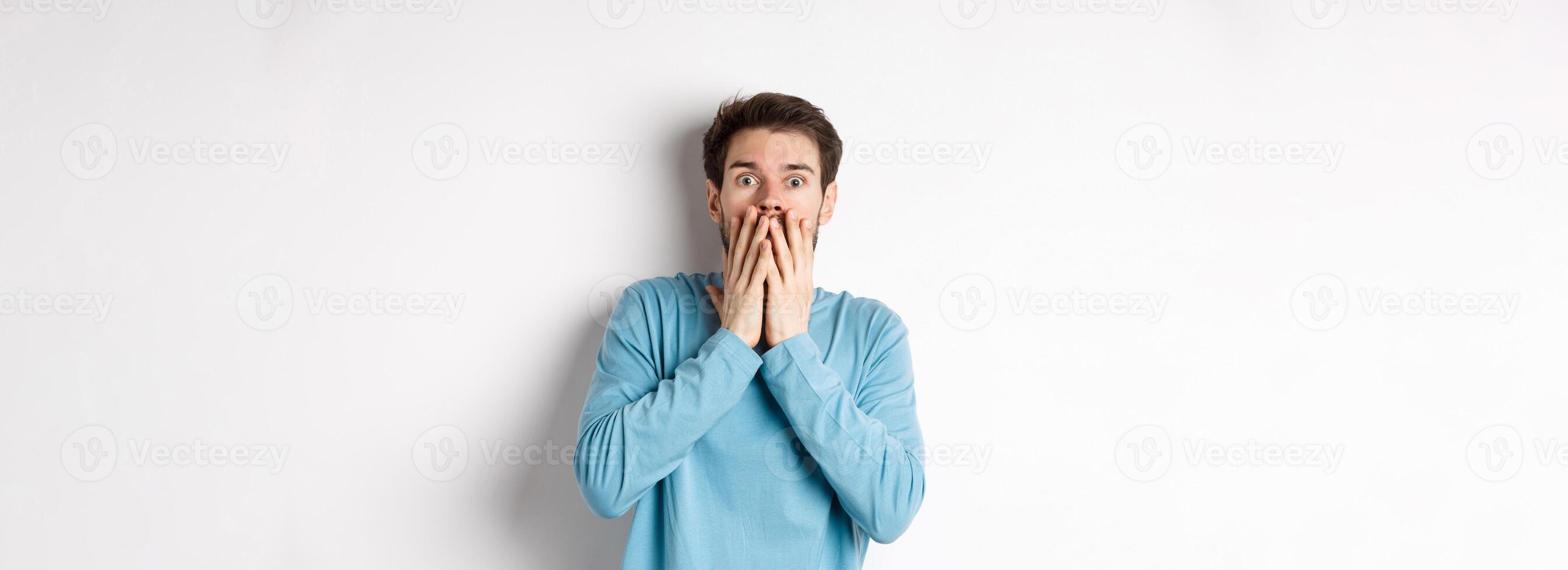 Shocked and startled cauacasian man, gasping and covering mouth with hands, staring at something scary, standing over white background photo