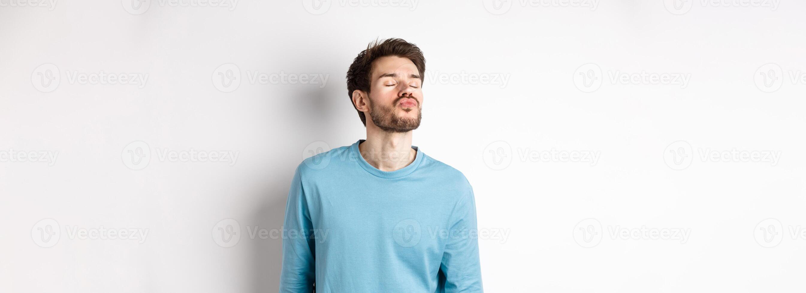 Image of cute and silly man waiting for kiss, pucker lips with closed eyes, standing over white background photo