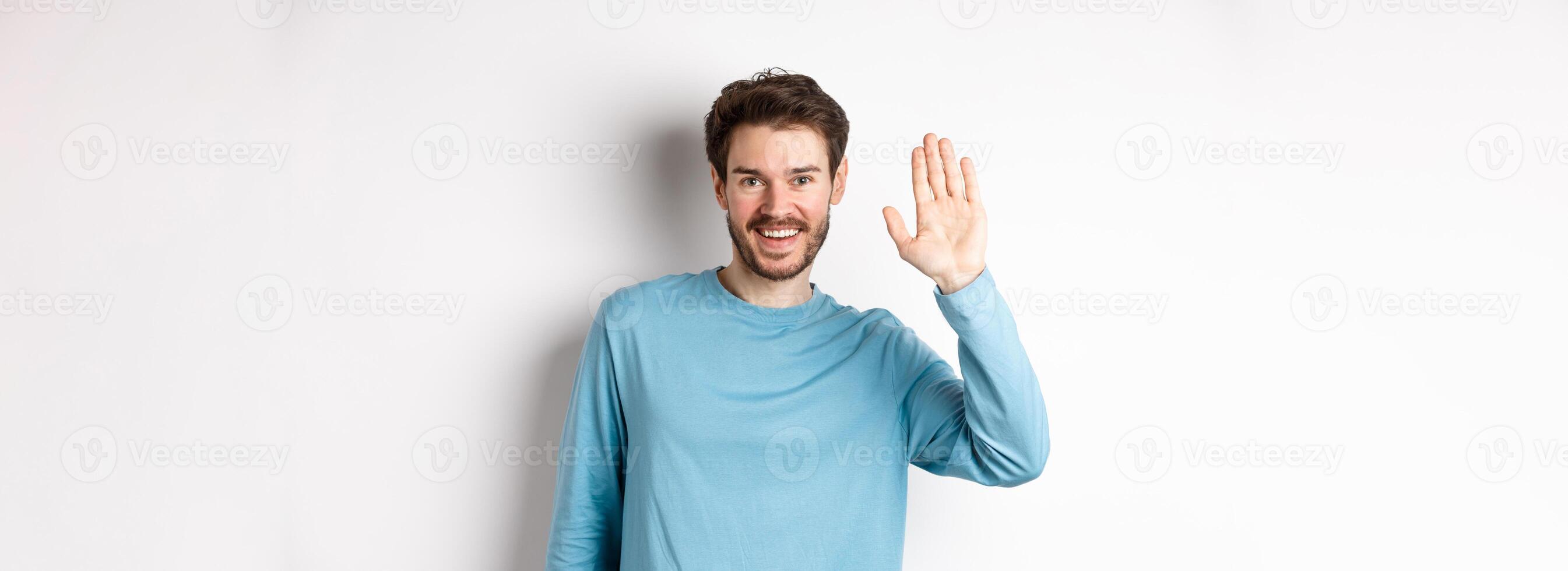 Cheerful young man with beard saying hello, looking friendly and waving hand to greet you, standing over white background photo