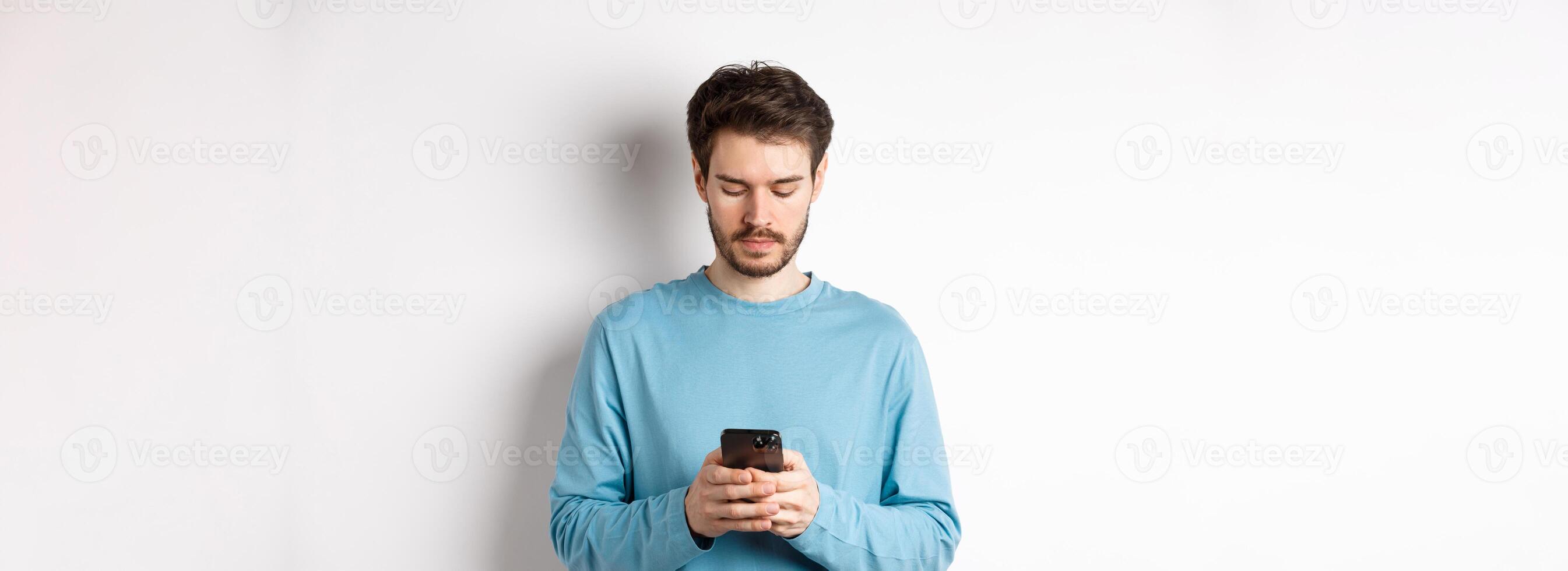Serious young man reading message on smartphone, looking at mobile screen, standing over white background photo