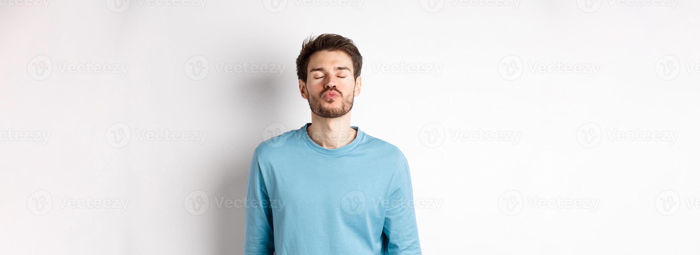 Attractive boyfriend waiting for kiss with puckered lips and closed eyes, standing in sweatshirt over white background photo