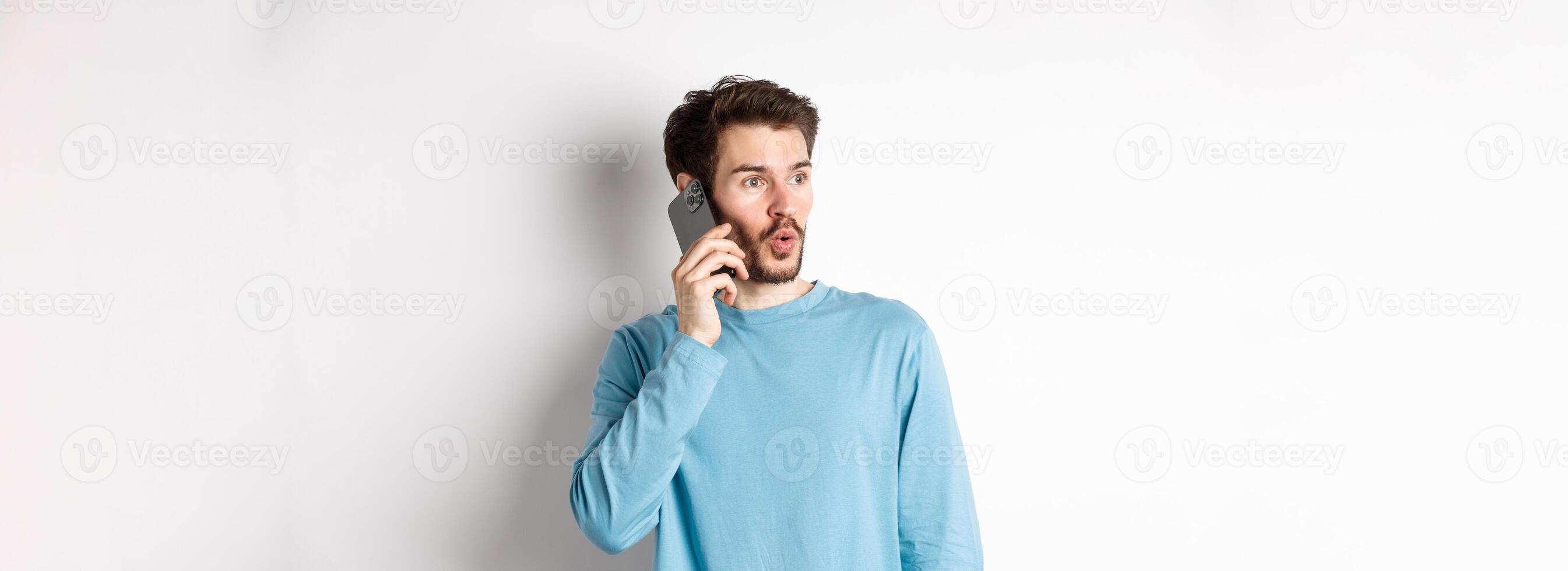 Technology concept. Intrigued young man looking left impressed, talking on mobile phone, calling someone, standing over white background photo