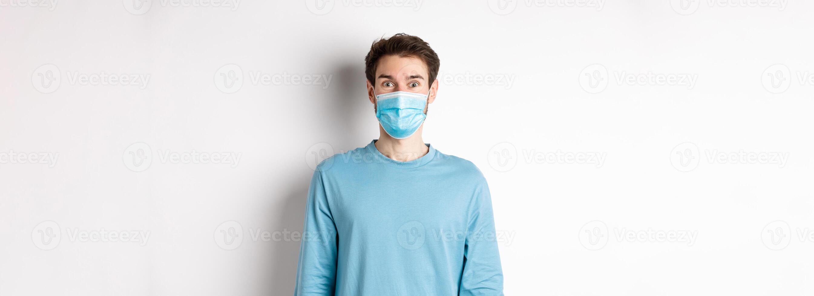 Covid-19, pandemic and social distancing concept. Cheerful man in medical mask raising eyebrows, looking surprised and excited at camera, standing over white background photo