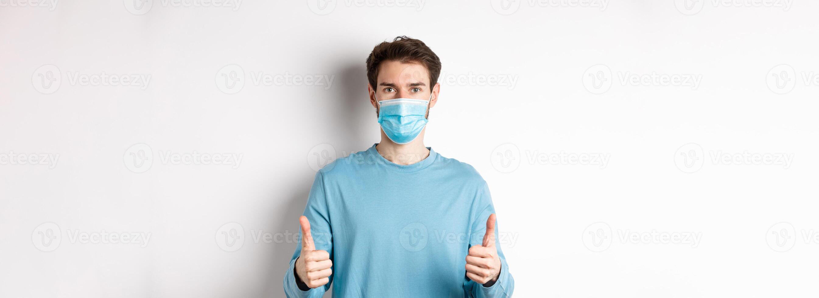 Covid-19, pandemic and social distancing concept. Young man in casual clothes and medical mask, showing thumbs up and looking serious, protecting from virus during quarantine photo
