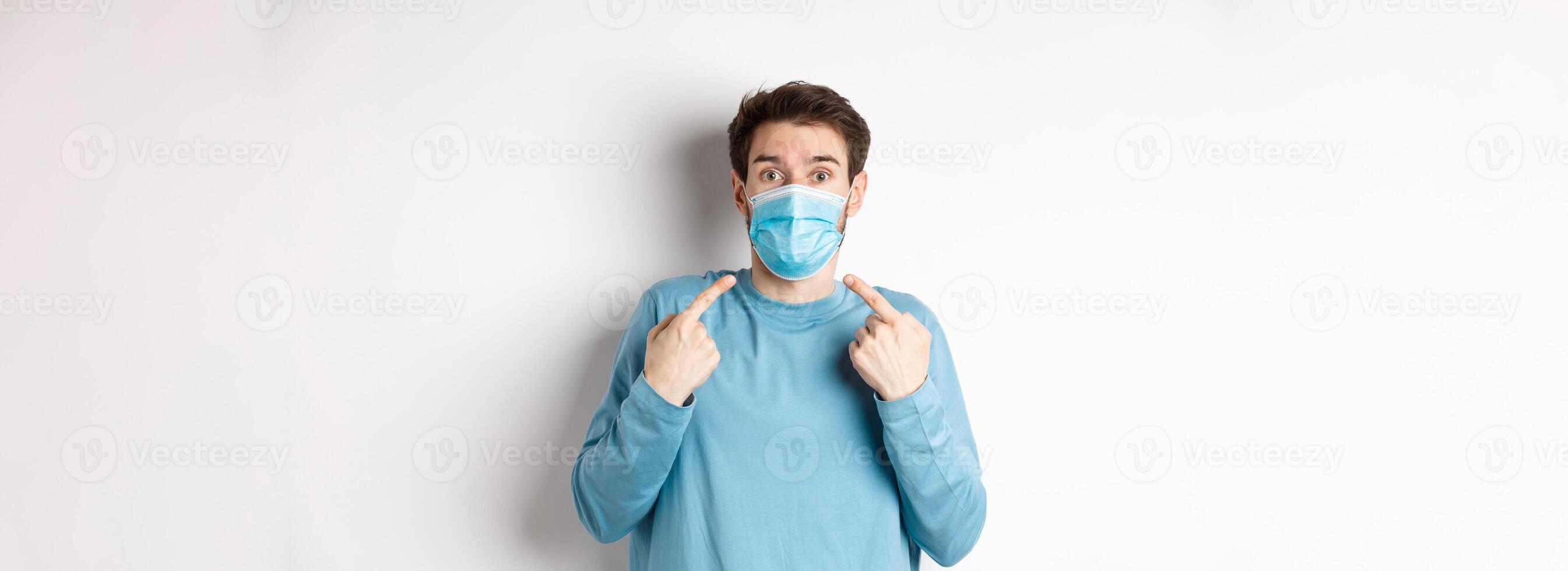 Coronavirus, health and quarantine concept. Confused guy pointing at his medical mask and looking surprised at camera, standing over white background photo