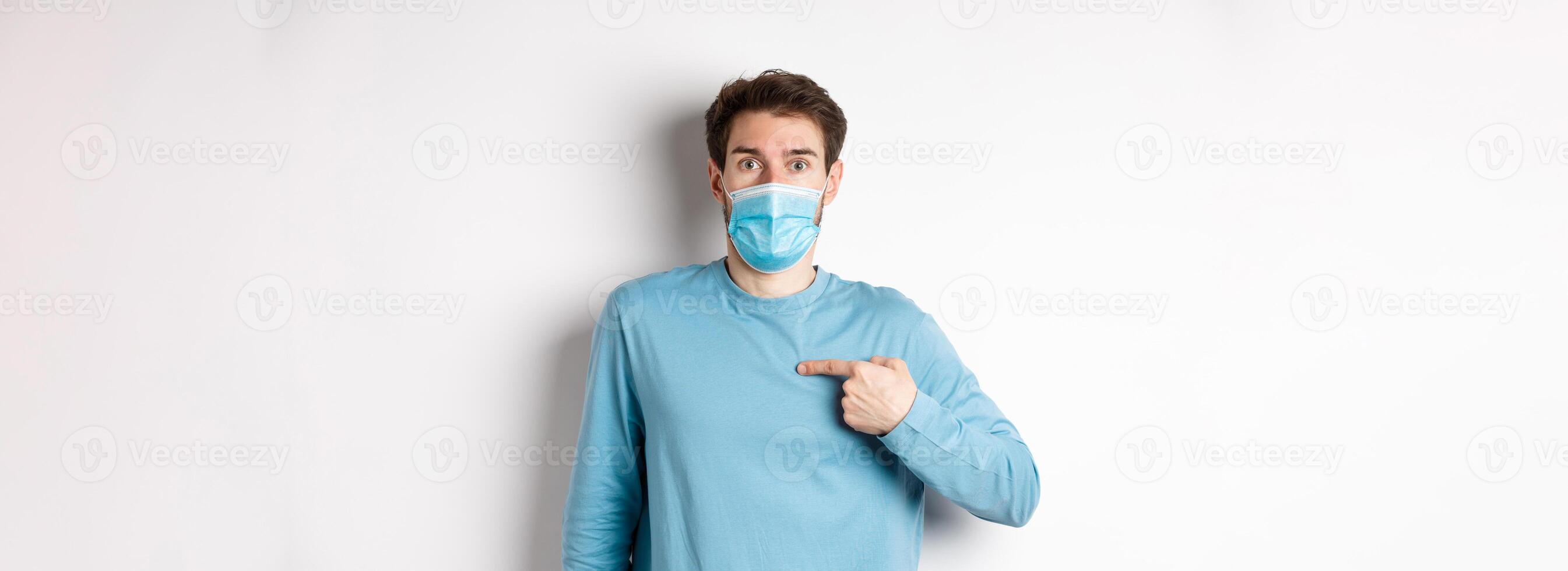 Coronavirus, health and quarantine concept. Surprised guy in medical mask points at himself, standing over white background photo