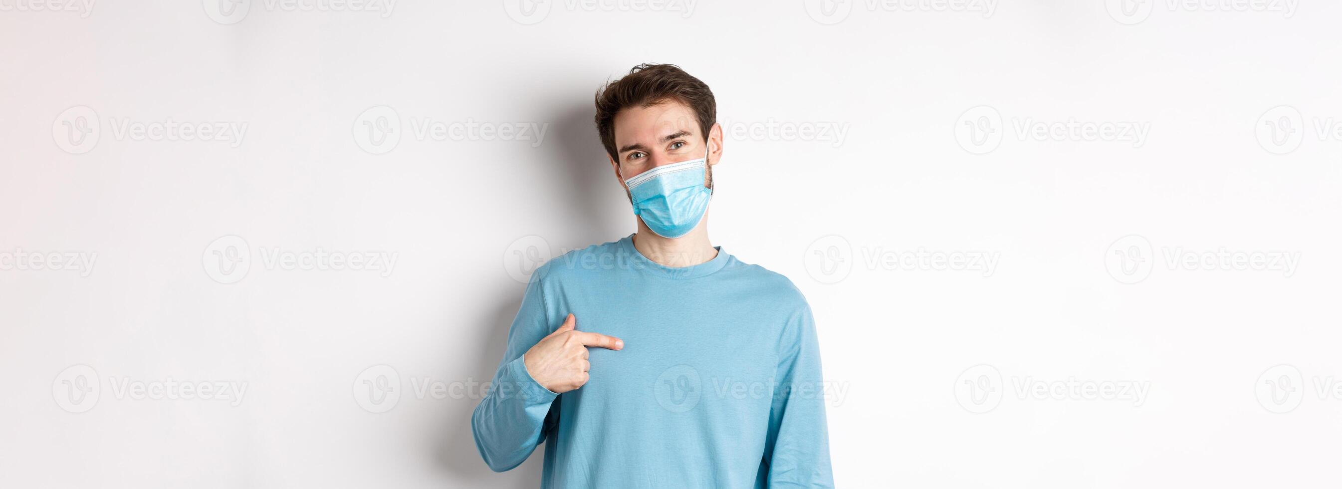 Covid-19, health and quarantine concept. Smiling caucasian man in medical mask pointing at himself, volunteering, standing over white background photo