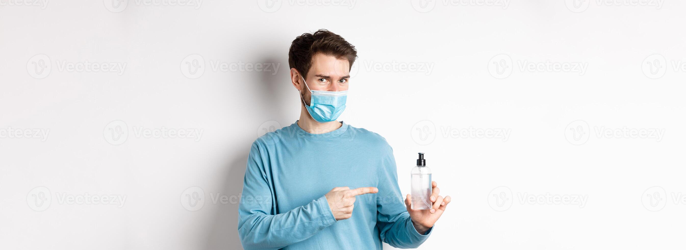 Covid-19, health and quarantine concept. Caucasian man in medical mask pointing finger at bottle with hand sanitizer, recommending antiseptic, standing on white background photo