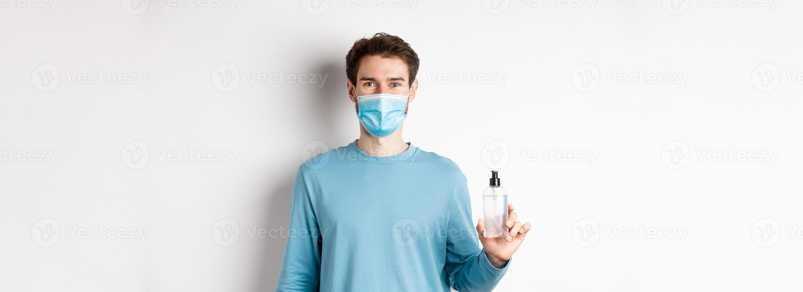 Covid-19, health and quarantine concept. Portrait of smiling man in medical mask showing hand sanitizer bottle, standing over white background photo