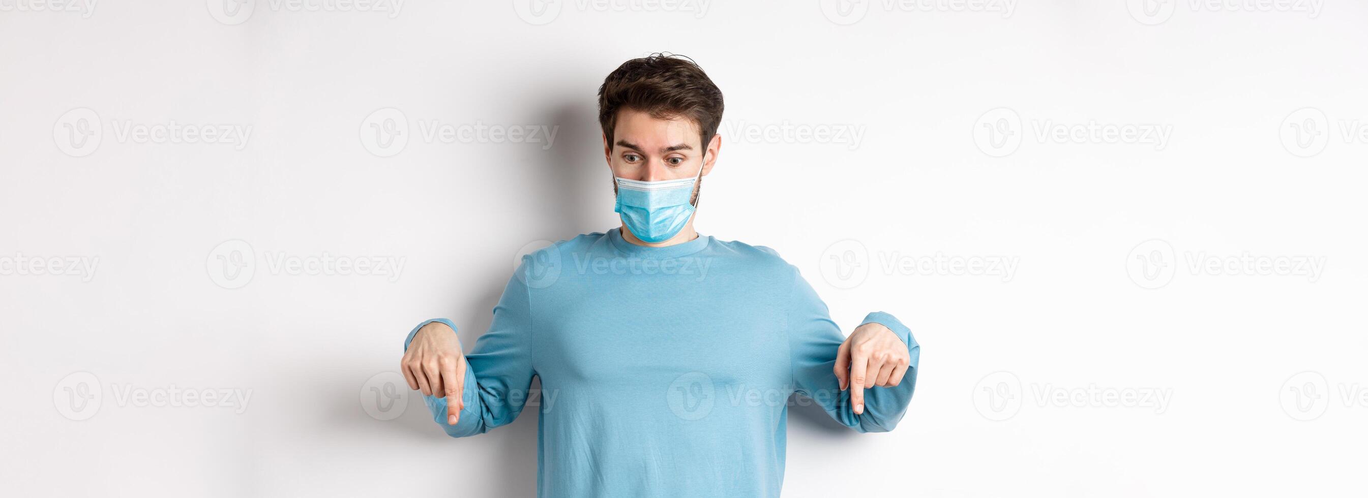 Covid-19, health and quarantine concept. Curious man checking out advertisement, pointing and looking down, wearing medical mask with casual blue sweatshirt, white background photo