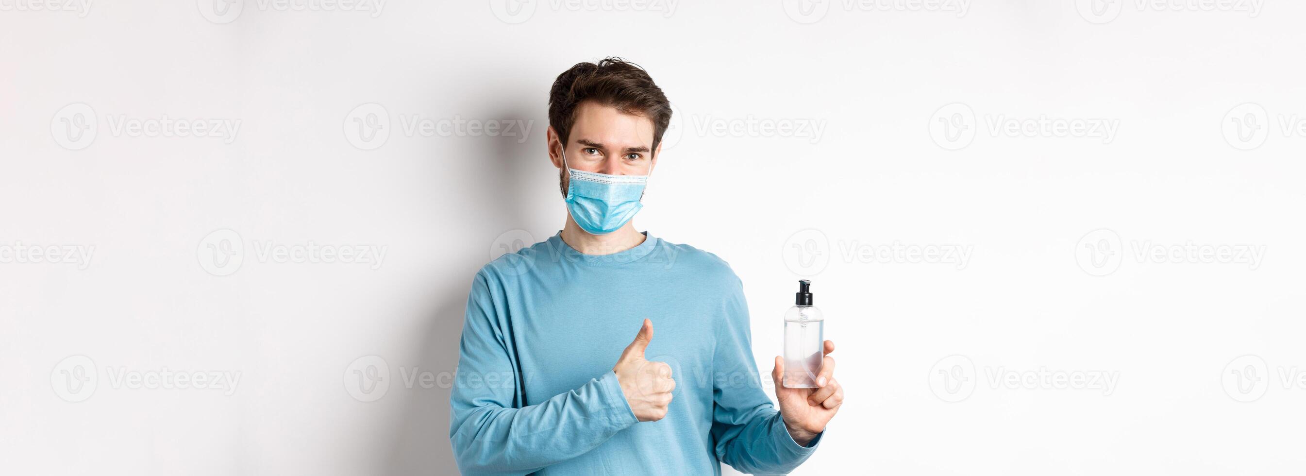 Covid-19, health and quarantine concept. Handsome smiling man in face mask, showing thumbs up and hand sanitizer bottle, recommending antiseptic brand, white background photo