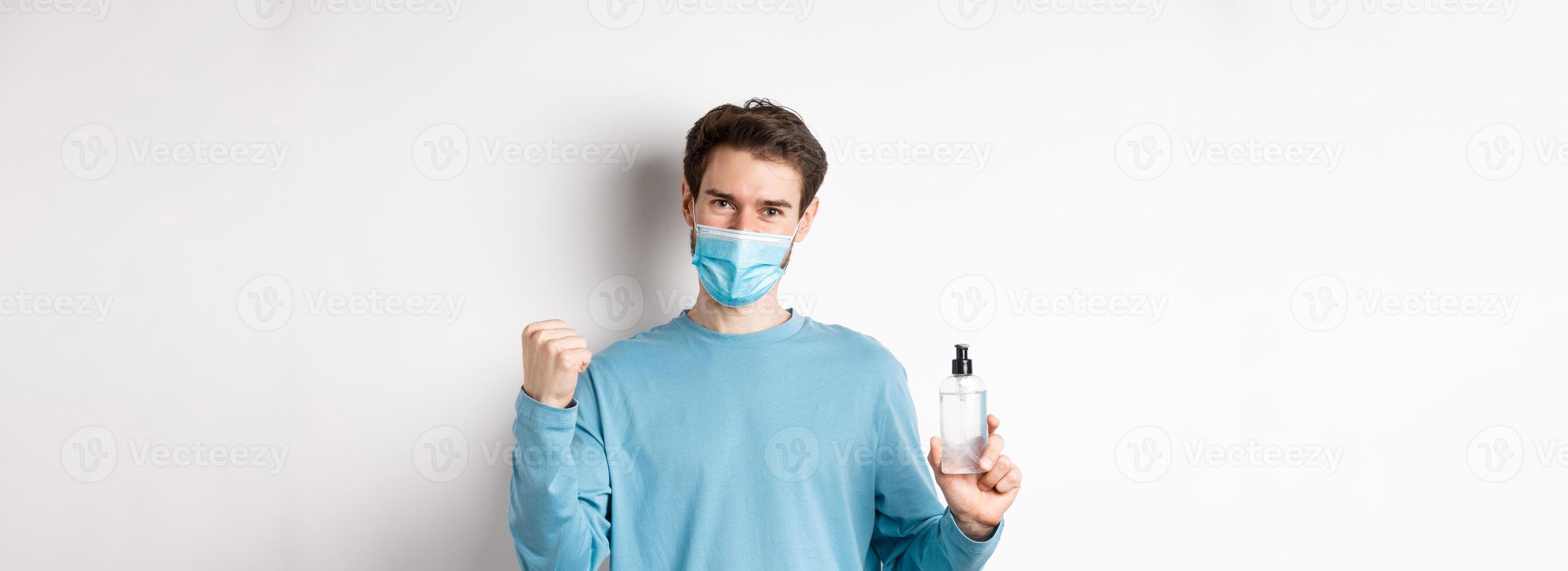 Covid-19, health and quarantine concept. Cheerful man in face mask celebrating, showing fist pump and bottle with hand sanitizer, fighting germs, white background photo