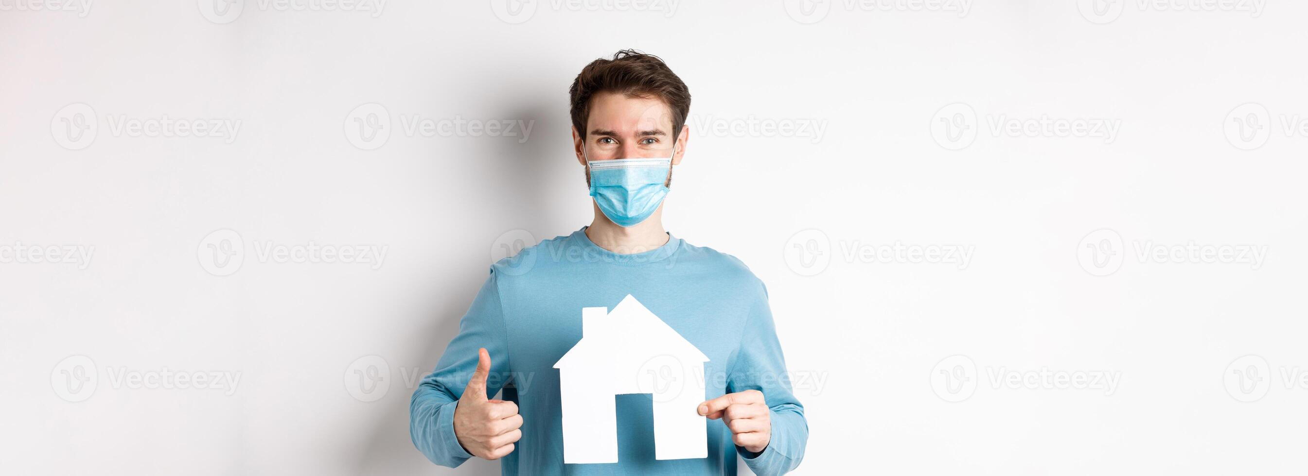 Covid and real estate concept. Smiling agency client showing thumb up and paper house cutout, wearing medical mask, approve good deal photo