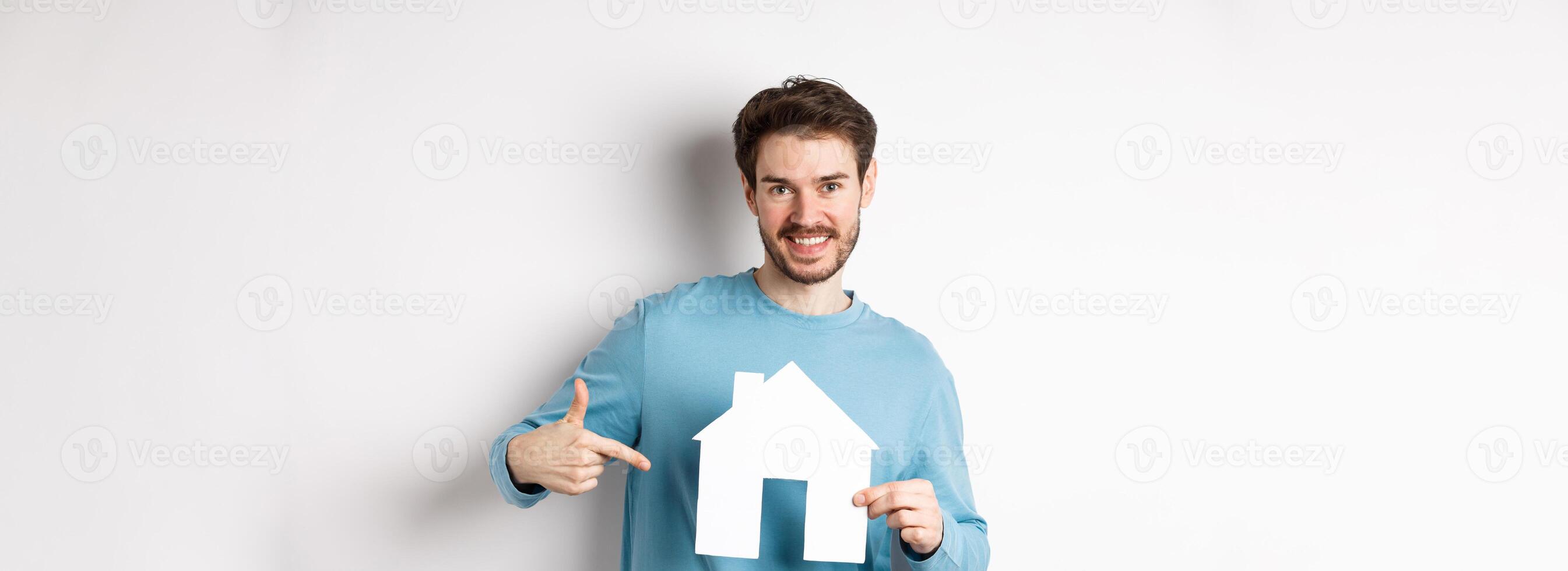 Real estate and insurance concept. Smiling young man pointing at paper house cutout and looking happy, buy apartment, standing over white background photo