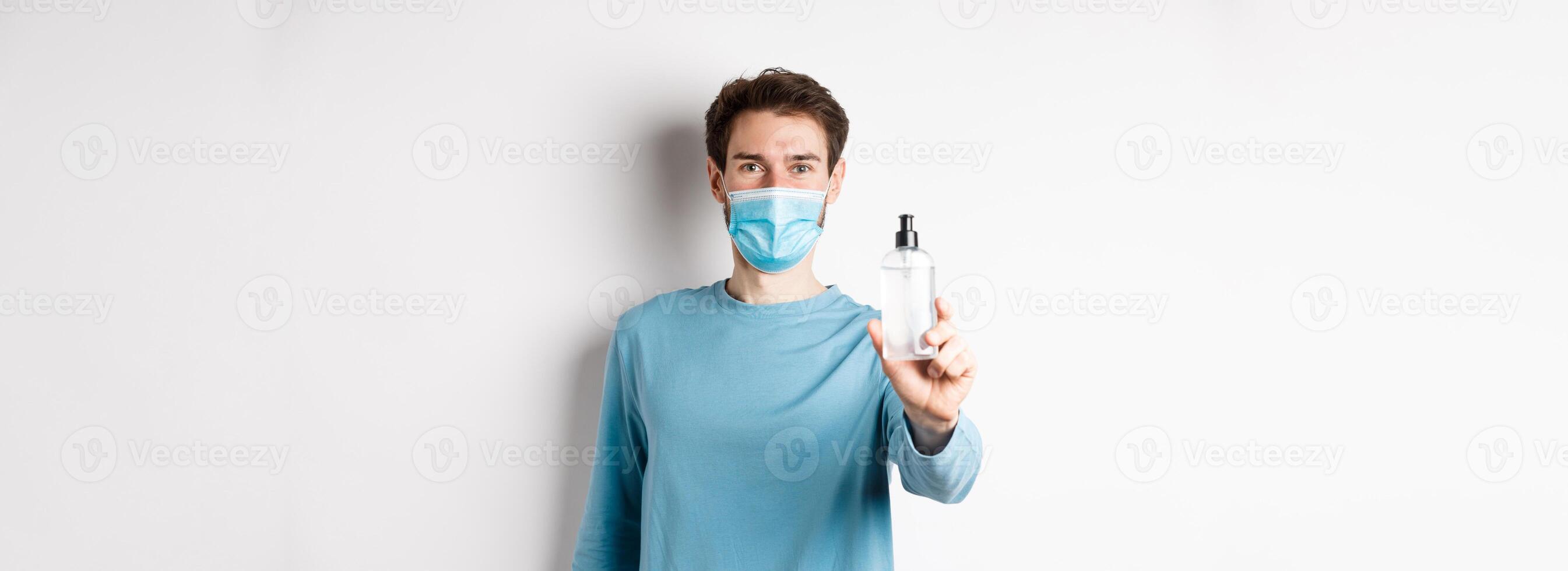 Covid-19, health and quarantine concept. Young man in medical mask stretch out hand and showing sanitizer, recommending antiseptic, standing over white background photo