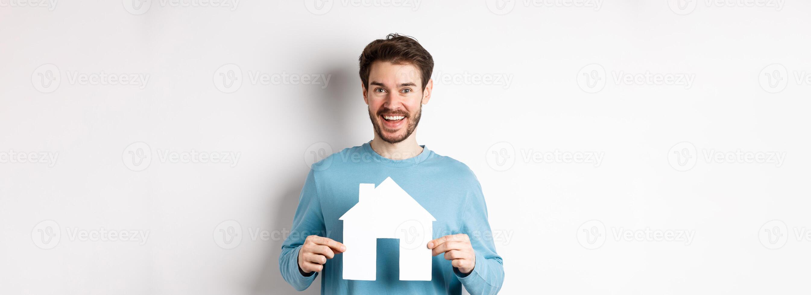 Real estate and insurance concept. Handsome modern man buying property, smiling and showing paper house cutout, standing over white background photo