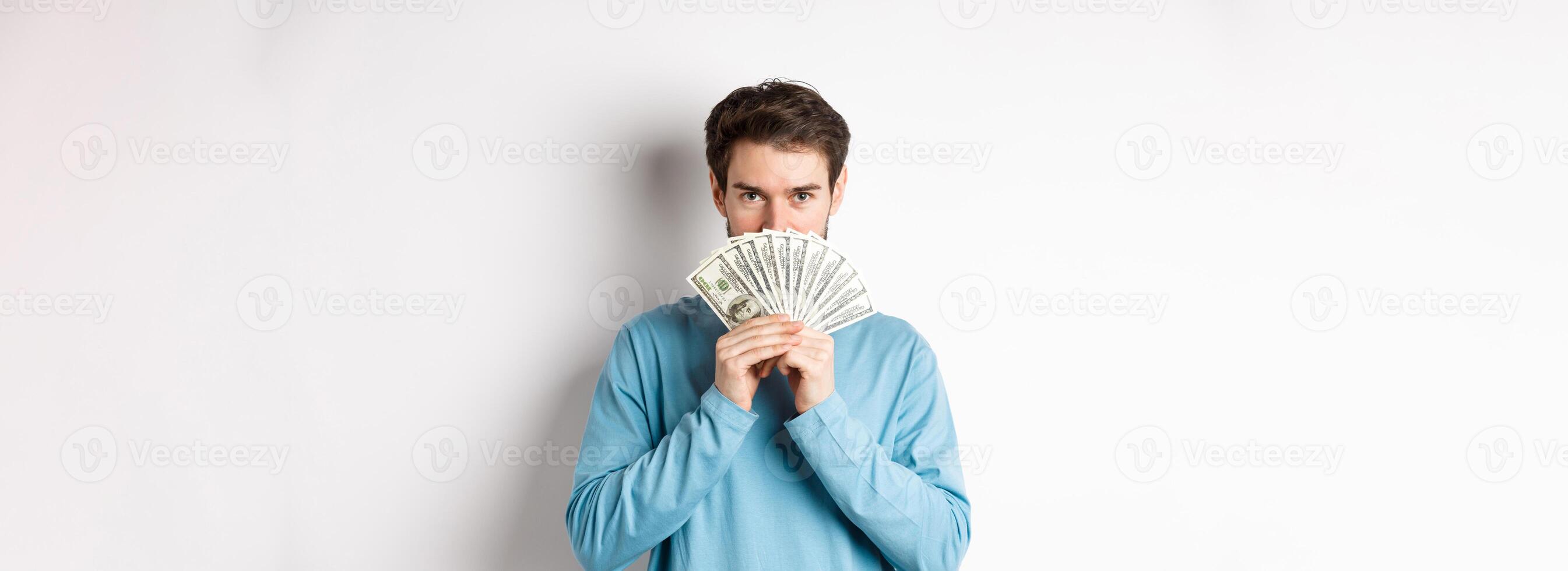 Finance concept. Smiling young man covering face behind money, showing dollars and looking happy, standing over white background photo