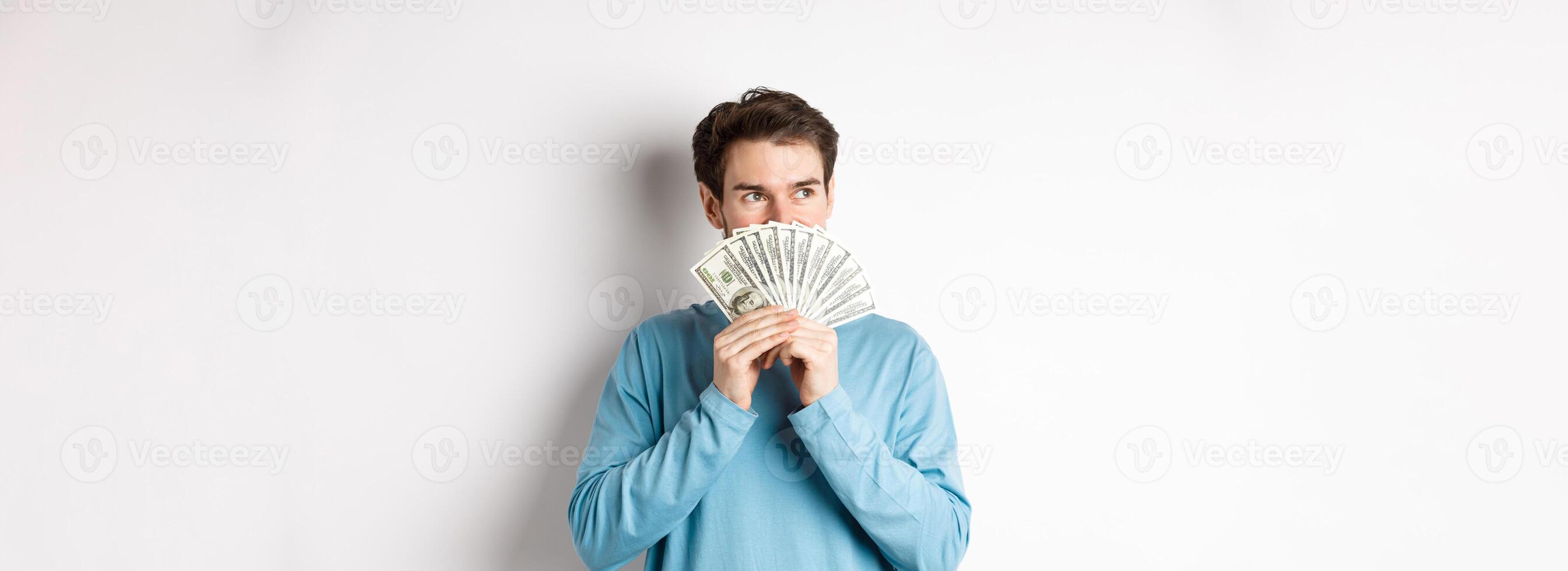 Dreamy man thinking of buying product, holding money and looking left pensive, imaging shopping, standing over white background photo