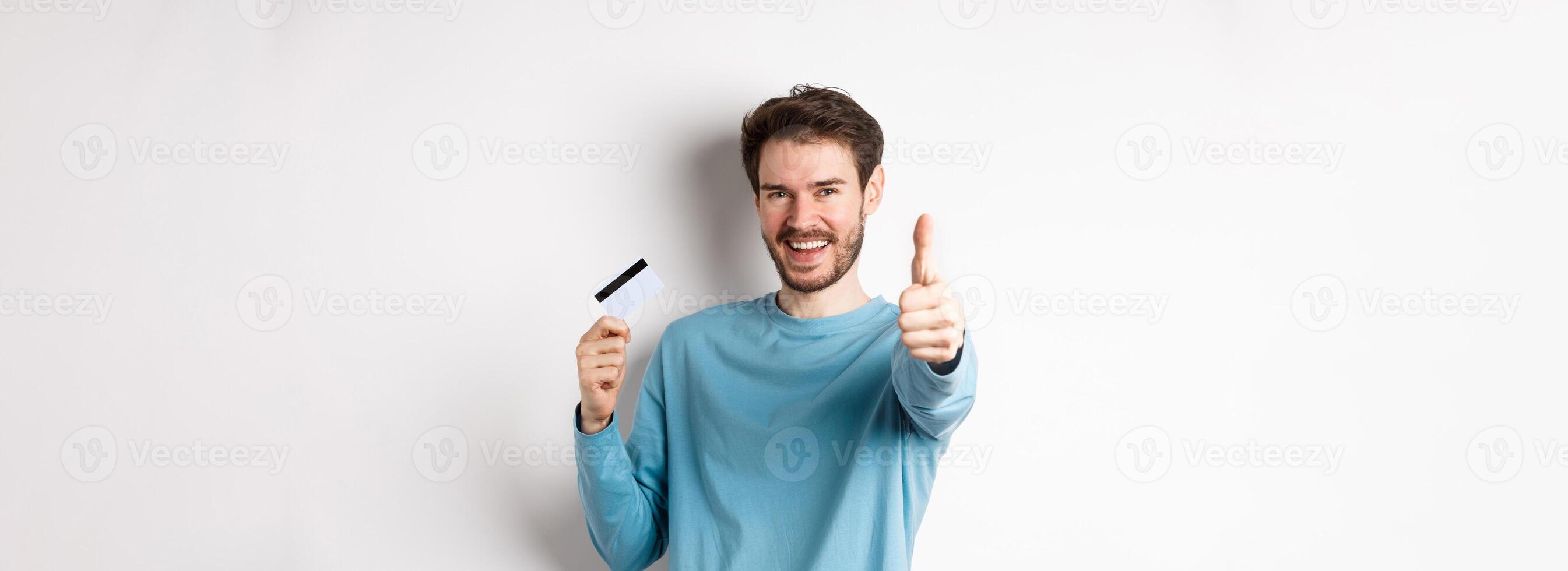 Cheerful young man showing plastic credit card and thumb up, like and approve good bank service, standing over white background photo