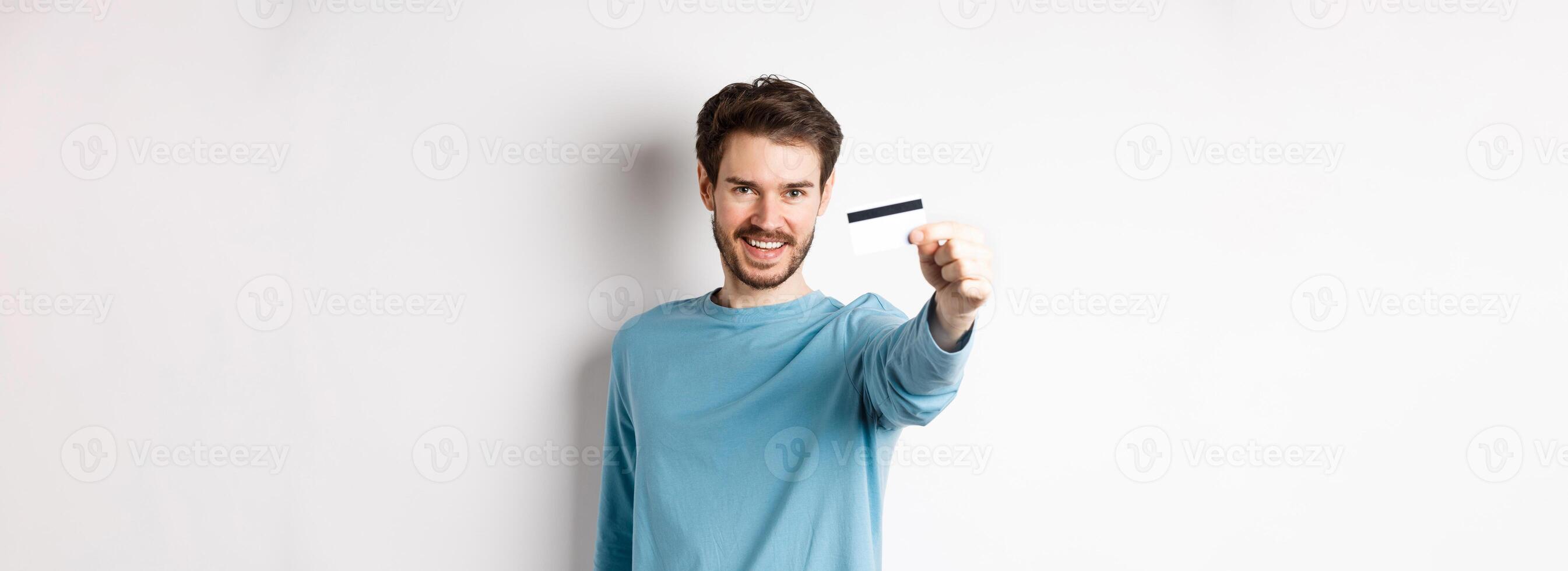 Confident young man smiling, stretch out hand and showing plastic credit card, standing on white background photo