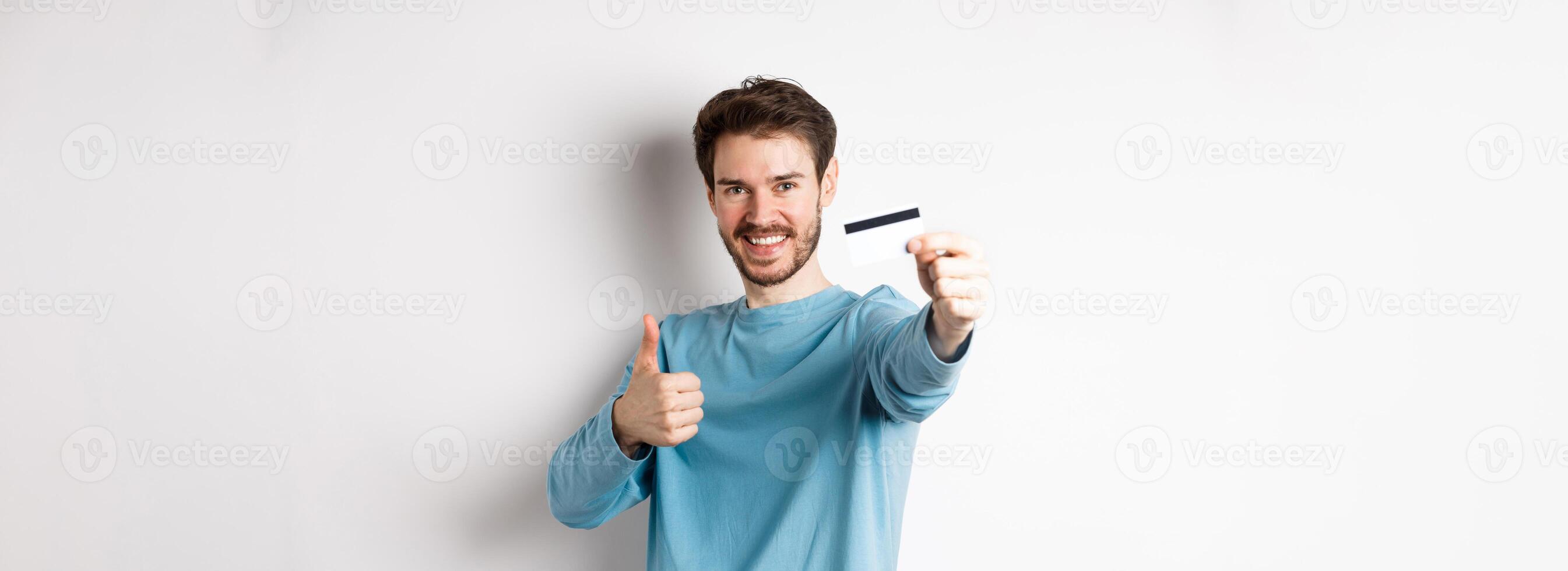 Handsome caucasian man showing plastic credit card with thumb-up, recommend and praise good bank offer, standing on white background. photo