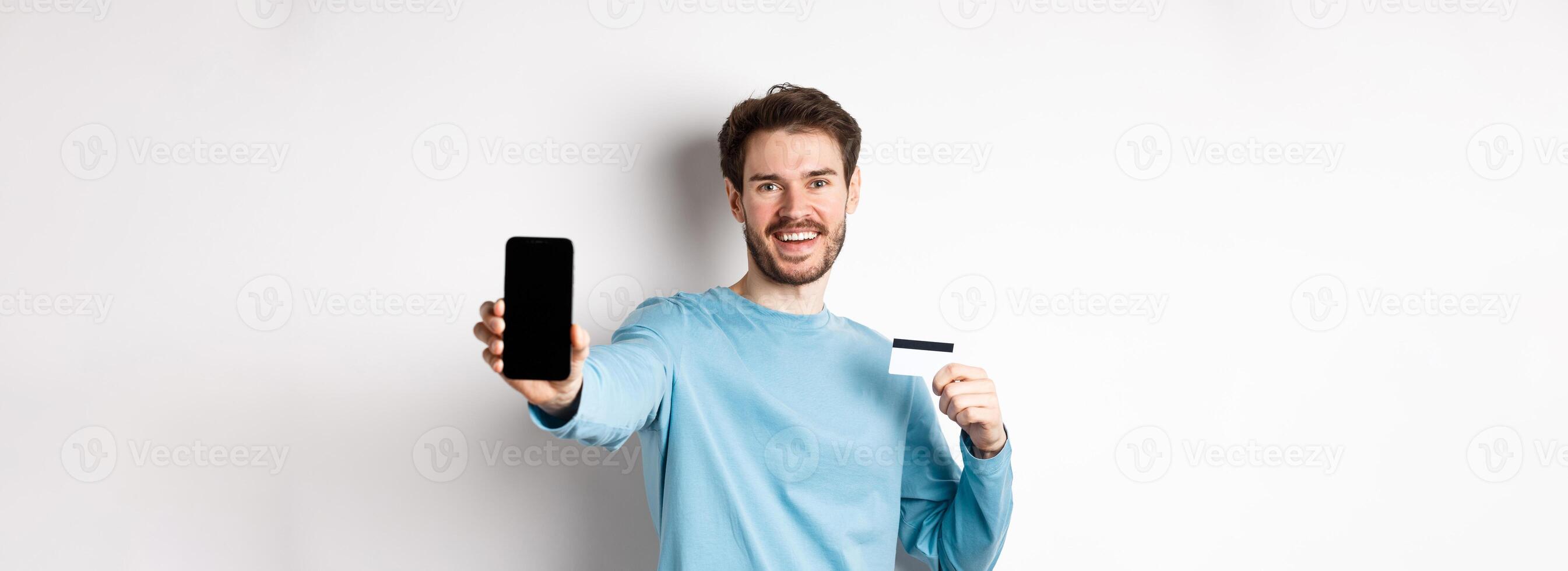 E-commerce and shopping concept. Smiling caucasian man showing plastic credit card and empty smartphone screen, recommending online app, white background photo