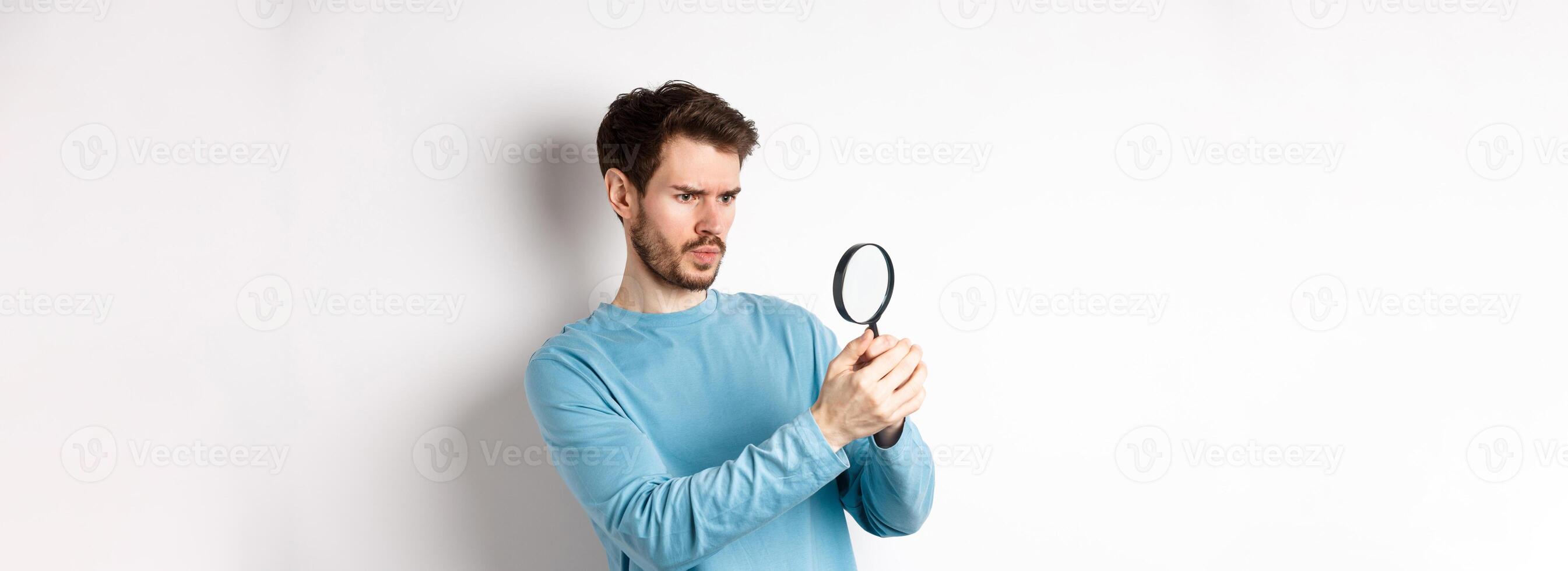 Serious-looking man looking through magnifying glass, investigating something, found interesting promo, standing on white background photo