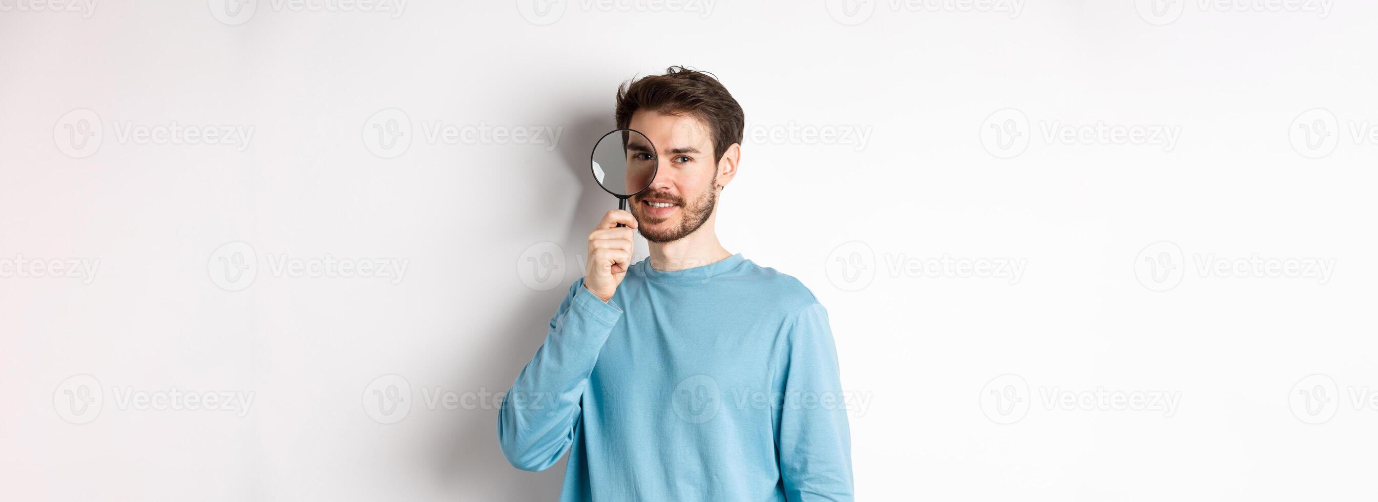 Smiling young man detective looking through magnifying glass, searching for promo offer, standing on white background photo
