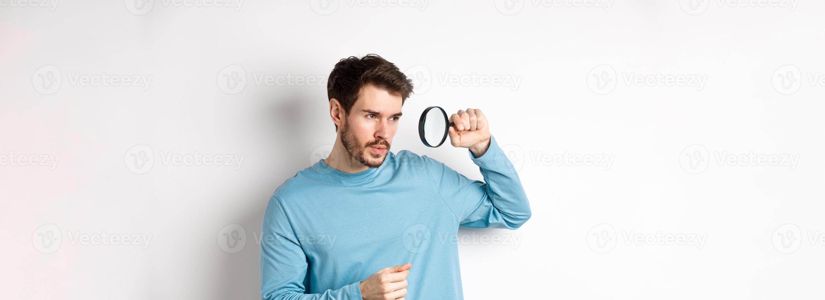 Young handsome man look through magnifying glass with curious face, investigating or searching for something, standing on white background photo