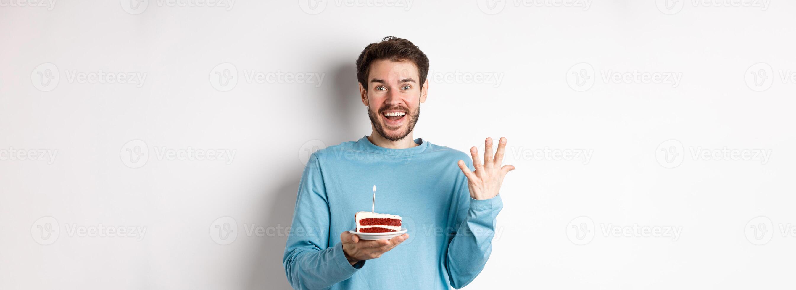 Excited man receive birthday surprise, holding bday cake and smiling happy, standing over white background, making wish on lit candle photo