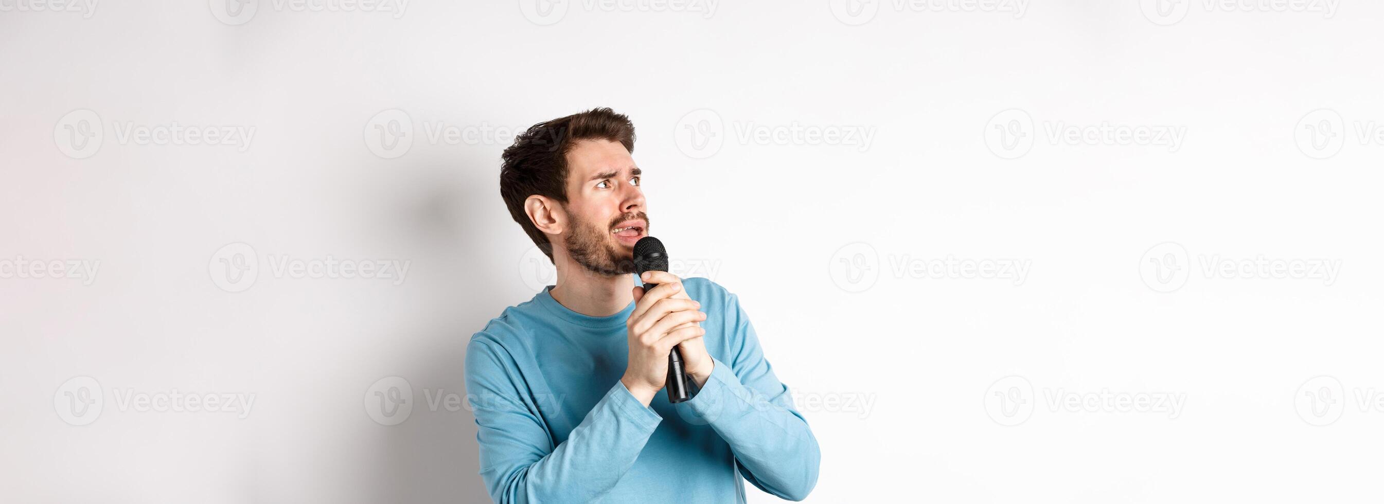 Worried and confused man reading lyrics on karaoke, looking left with unsure face, holding microphone and singing, white background photo
