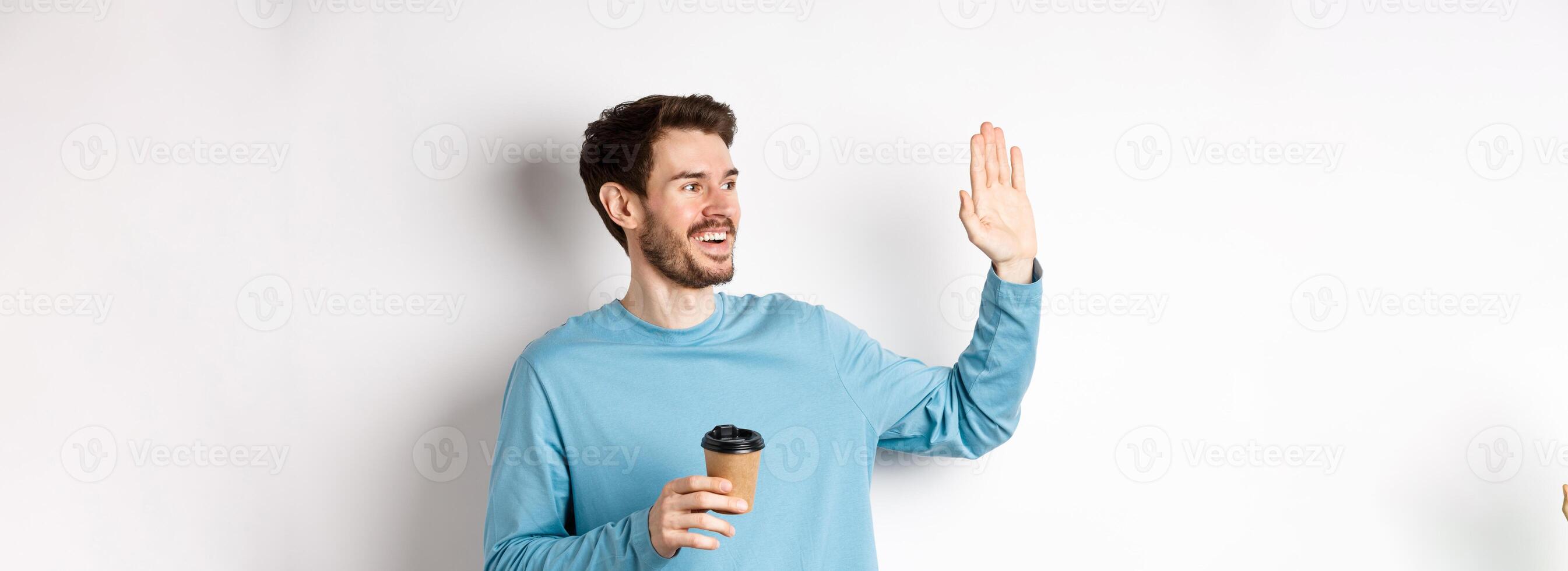 Friendly young man drinking coffee from takeaway cup and saying hello, waving hand left and smiling, standing over white background photo