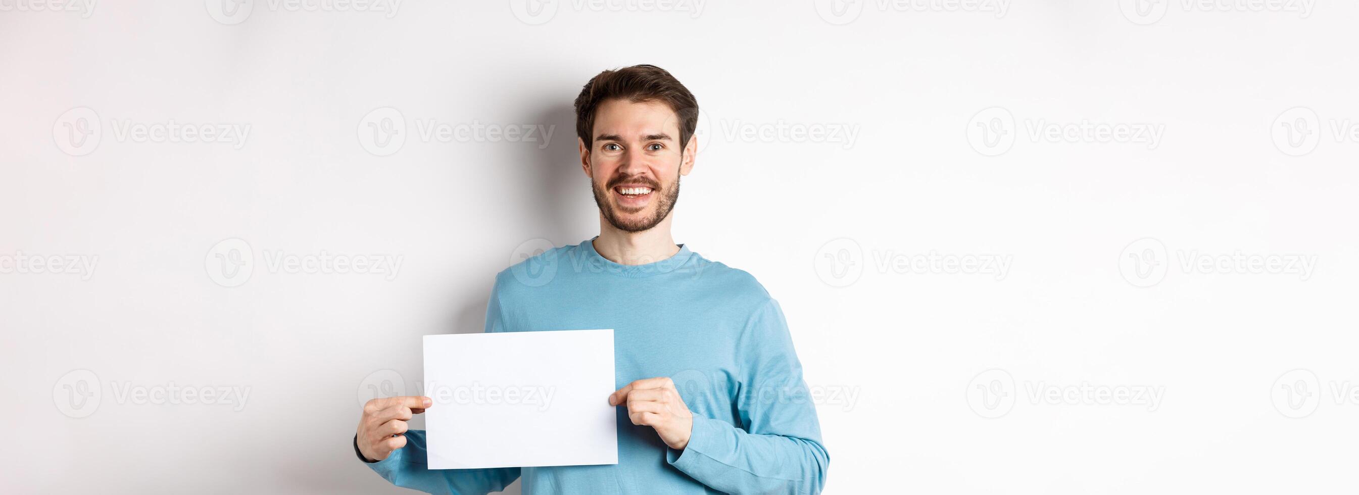 Young man with happy face showing blank piece of paper for your logo or sign, smiling at camera, standing over white background photo