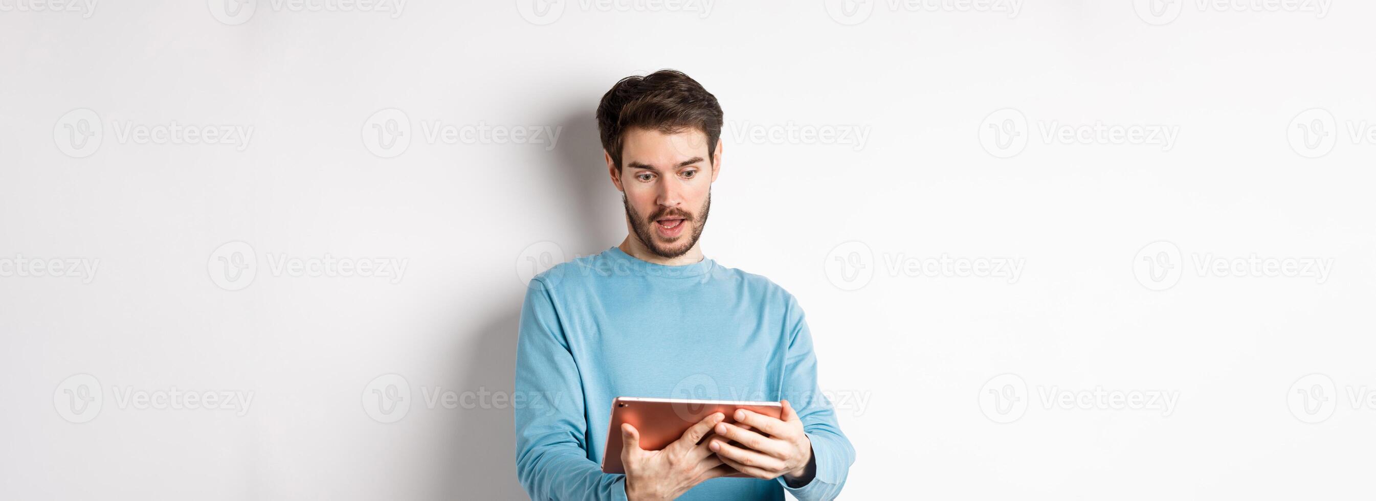 Image of man looking curious at digital tablet, reading interesting news online, standing on white background photo