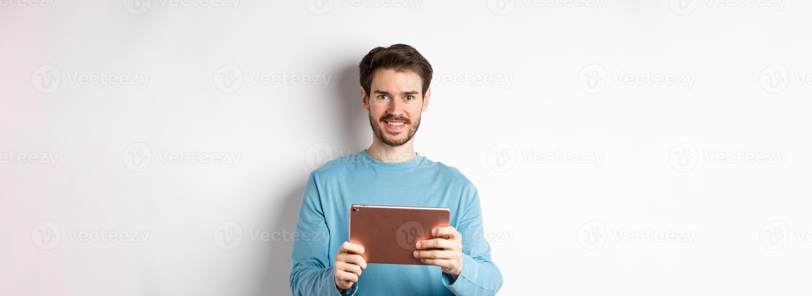 Handsome young man holding digital tablet and smiling, networking with device, standing over white background photo