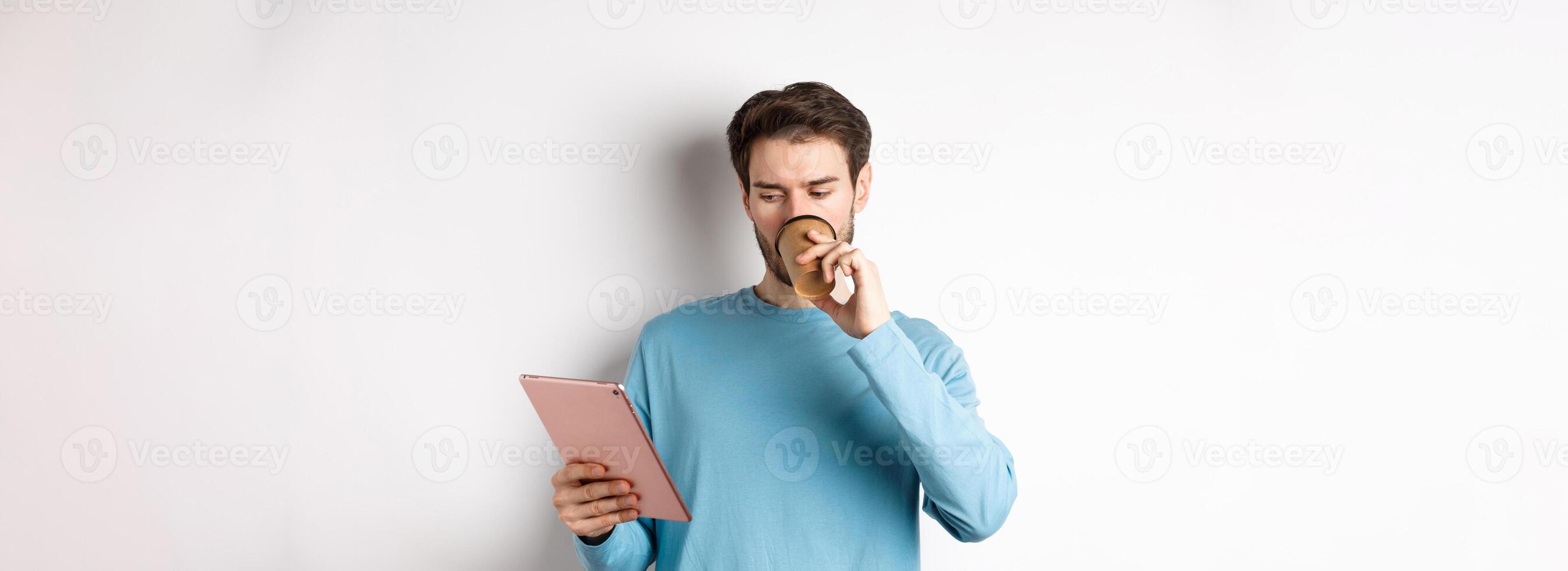 Handsome caucasian man drinking coffee and reading digital tablet screen, standing in blue sweatshirt over white background photo
