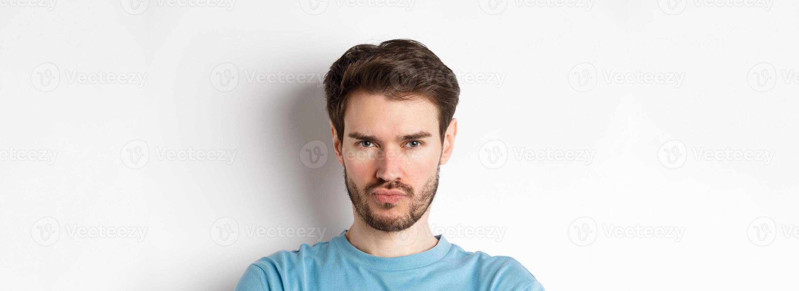 Close-up of grumpy young man grimacing, sulking and making offended face, standing upset over white background photo