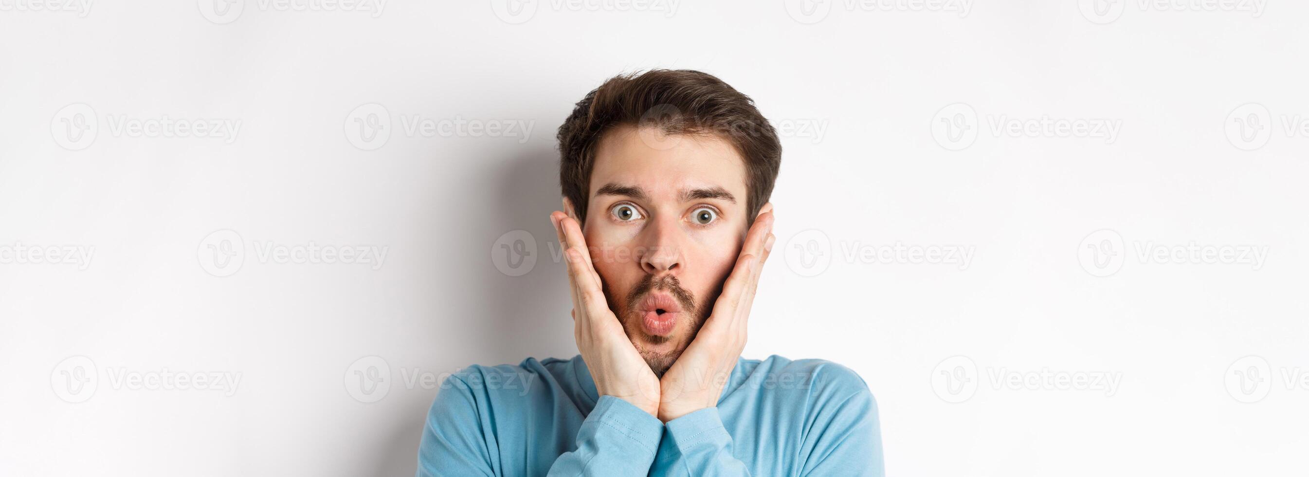 Close-up of impressed man checking out special offer, saying wow and touching face with amazed expression, stare at camera in awe, standing over white background photo