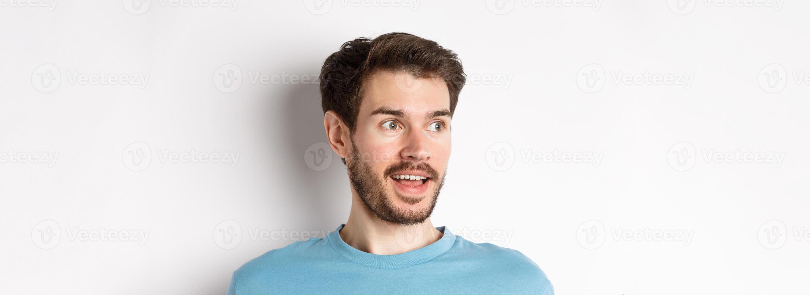 Close-up of surprised and excited bearded man, looking left with opened mouth and amazed face, checking out promo, white background photo