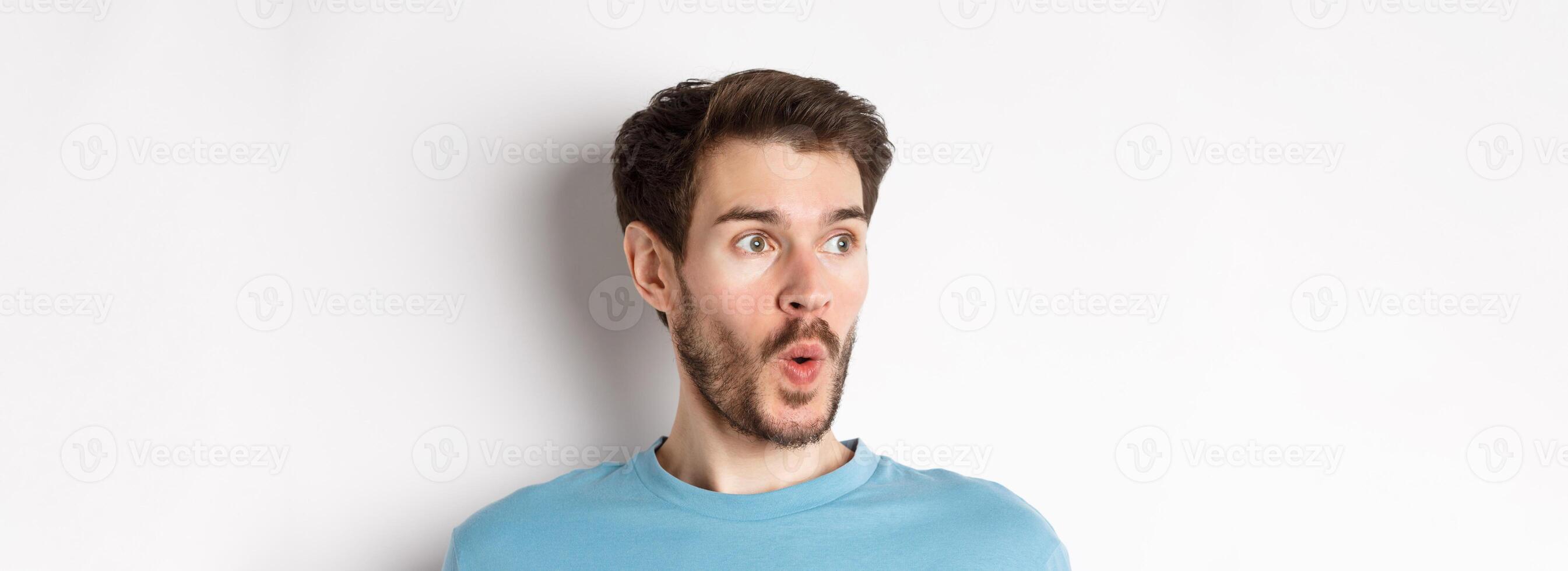Close-up of impressed caucasian man saying wow, looking left amazed, checking out promotion deal, white background photo