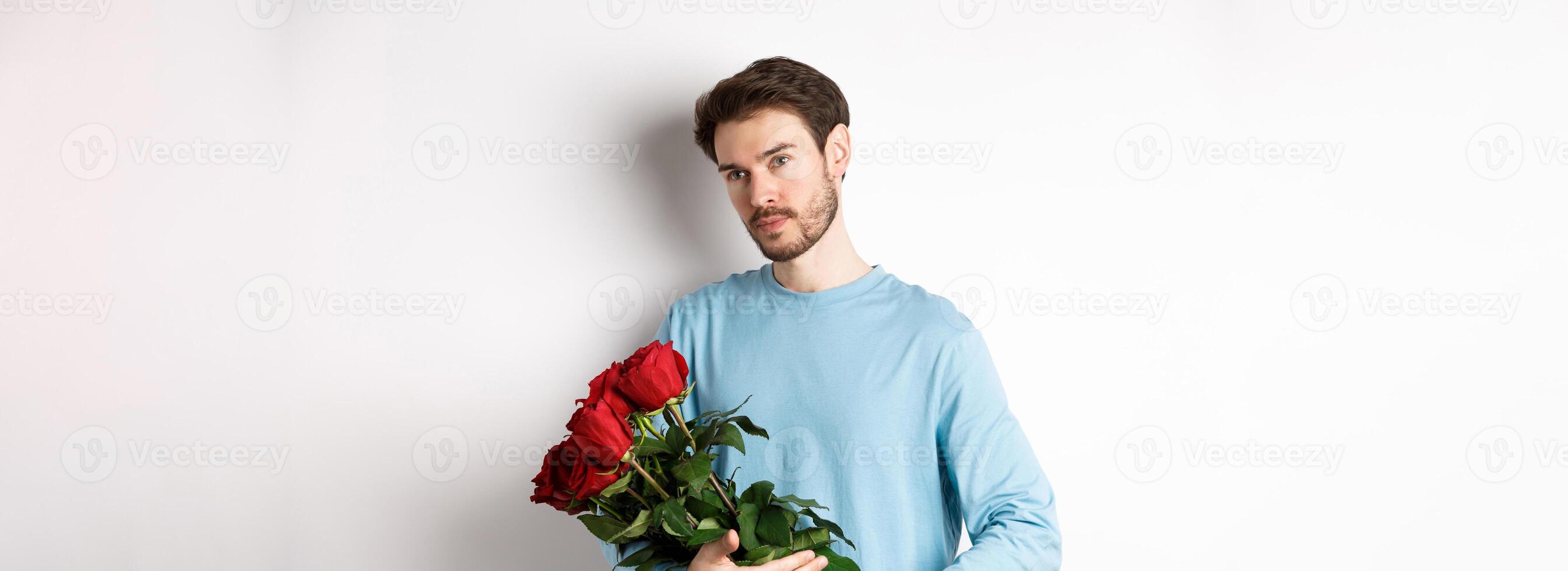 Handsome young man holding beautiful red roses for his lover on Valentines day, looking pensive, standing over white background photo