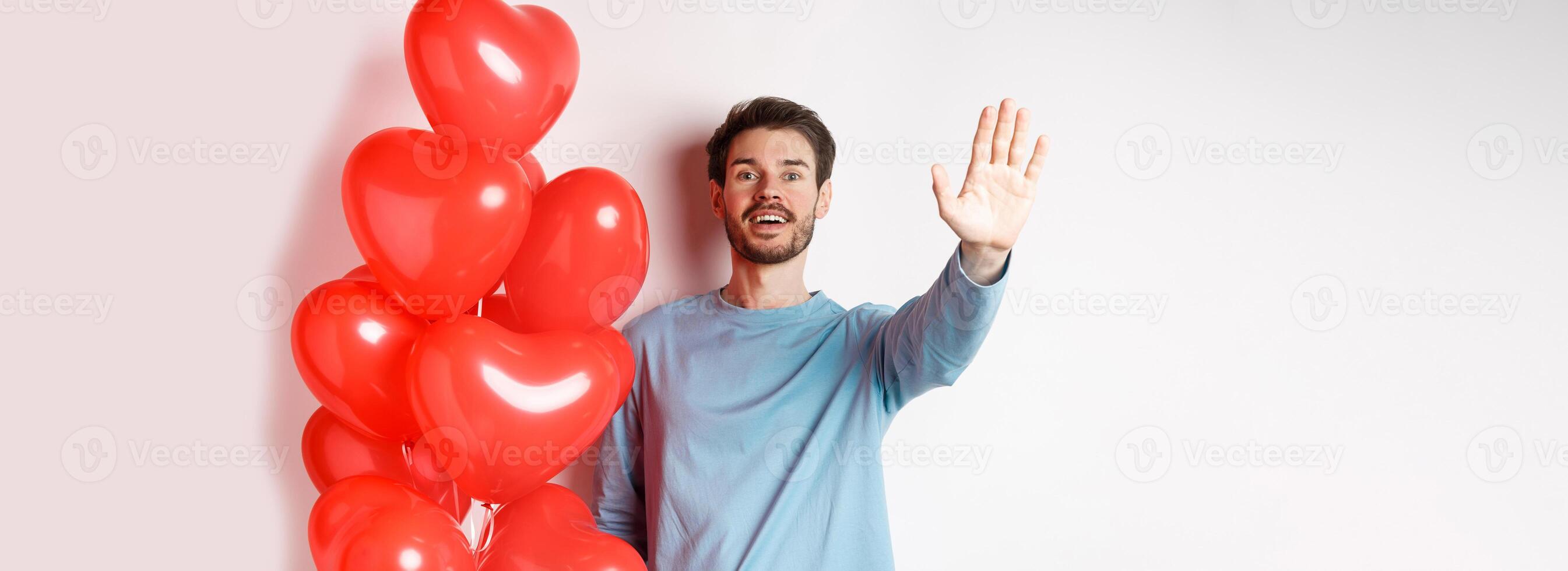 Handsome young caucasian man standing with romantic heart balloon and waving hand at lover, waiting for his date on Valentines day with cute surprise, standing over white background photo