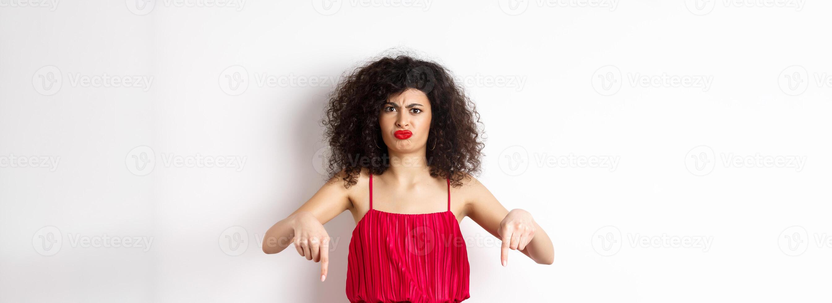 Disappointed caucasian woman in red dress complaining on spoiled date, frowning and grimacing, standing over white background photo