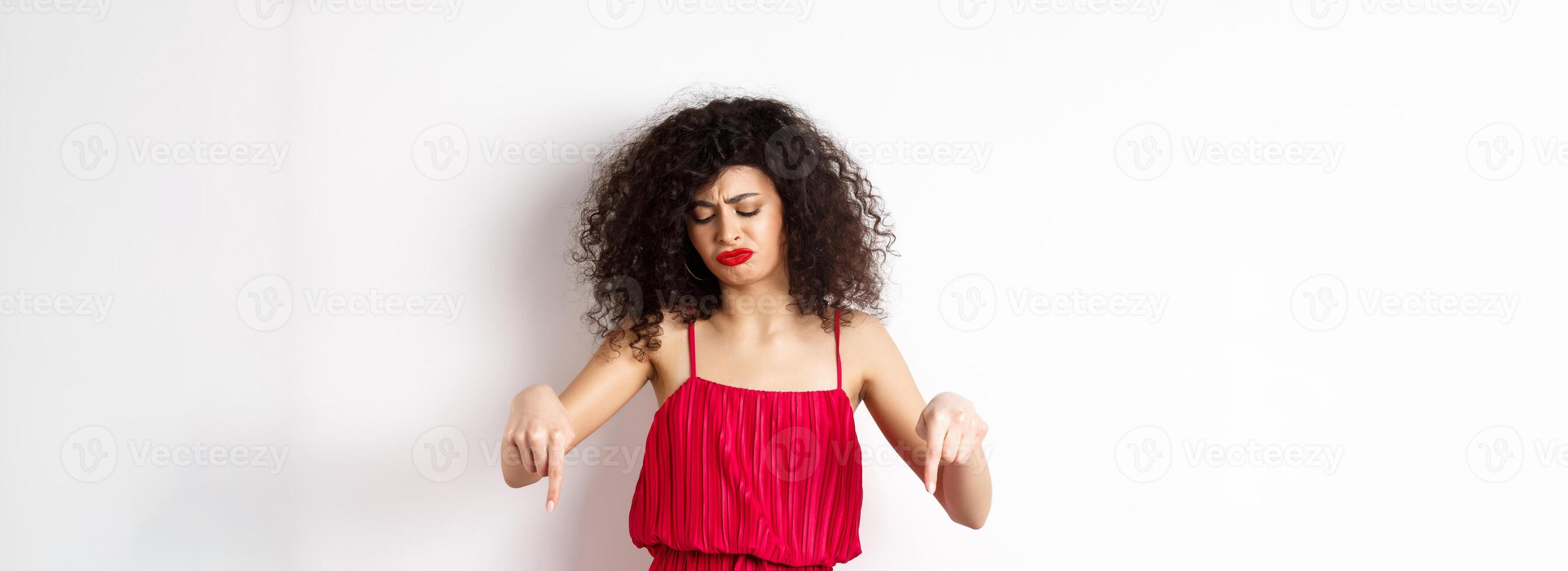Sad and disappointed curly-haired woman in red dress, frowning and looking down jealous, pointing at promo, standing over white background photo