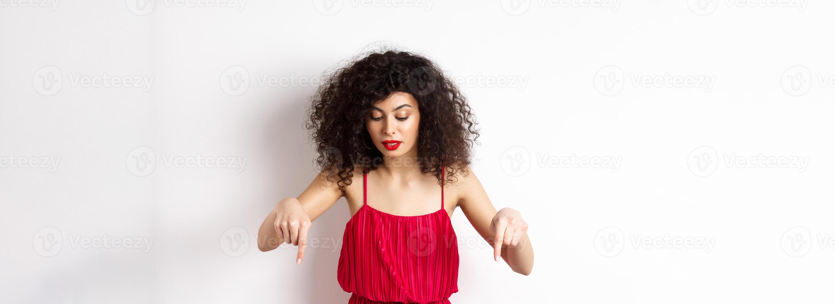 Intrigued beautiful woman with red lips and dress, pointing and looking down at banner, reading logo, standing over white background photo